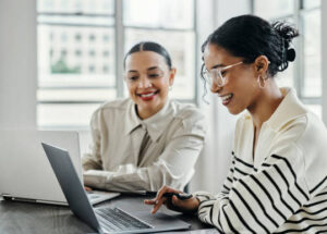 istockphoto-1438634414-612x612-1 Business women, laptop and and happy team in office for web design, collaboration and training. Planning, digital marketing and coaching by woman leader menor help, explain and support creative goal